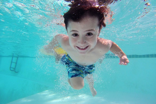 In einem Schwimmbecken unter Wasser sieht man ein Kind tauchen und dabei zeigt es lächelnd seine Zähne.