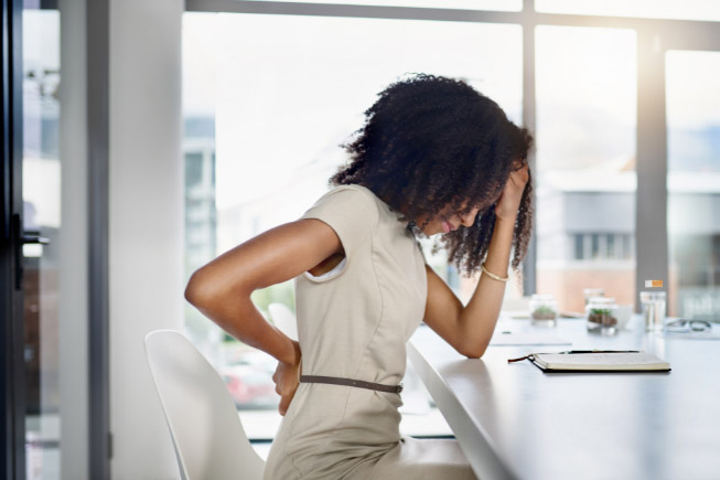Eine Frau sitzt an ihrem Schreibtisch und hält sich mit schmerzverzerrtem Gesicht den Rücken